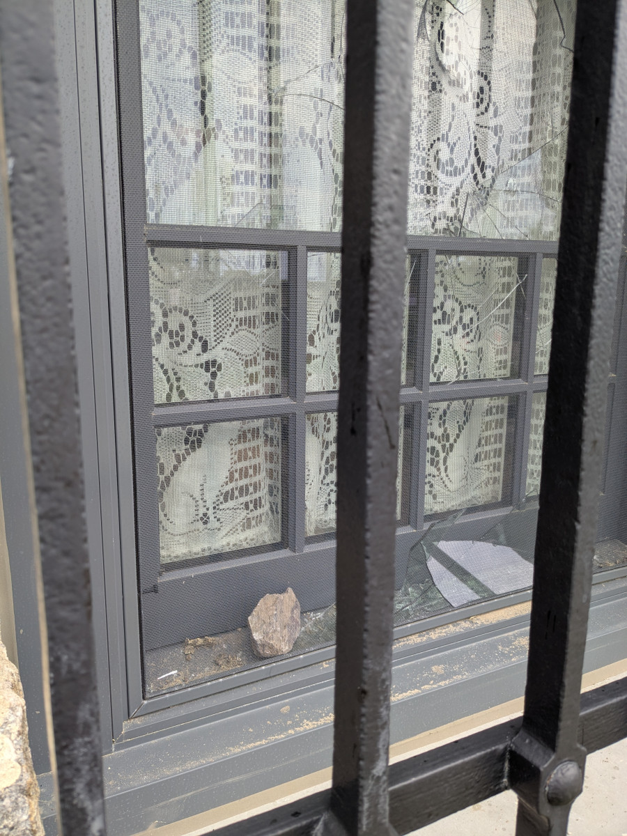 Shattered window glass viewed through iron security bars, with a lace curtain visible inside